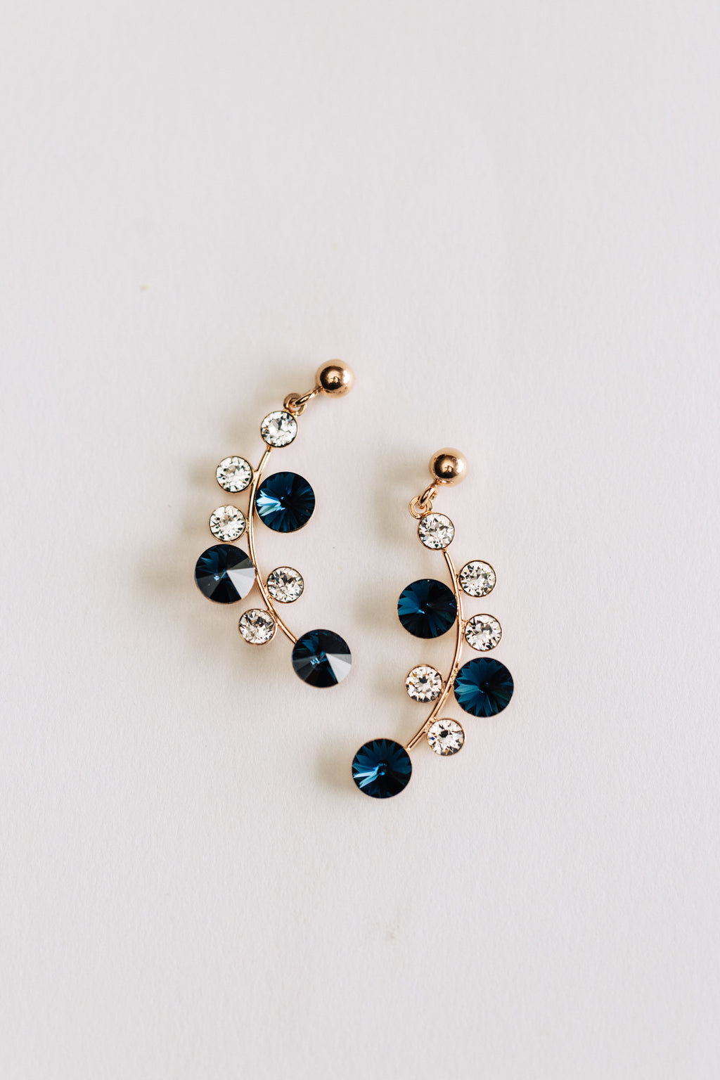 A pair of hypoallergenic rose gold earrings featuring blue and clear crystals arranged in a circular pattern on a white background.