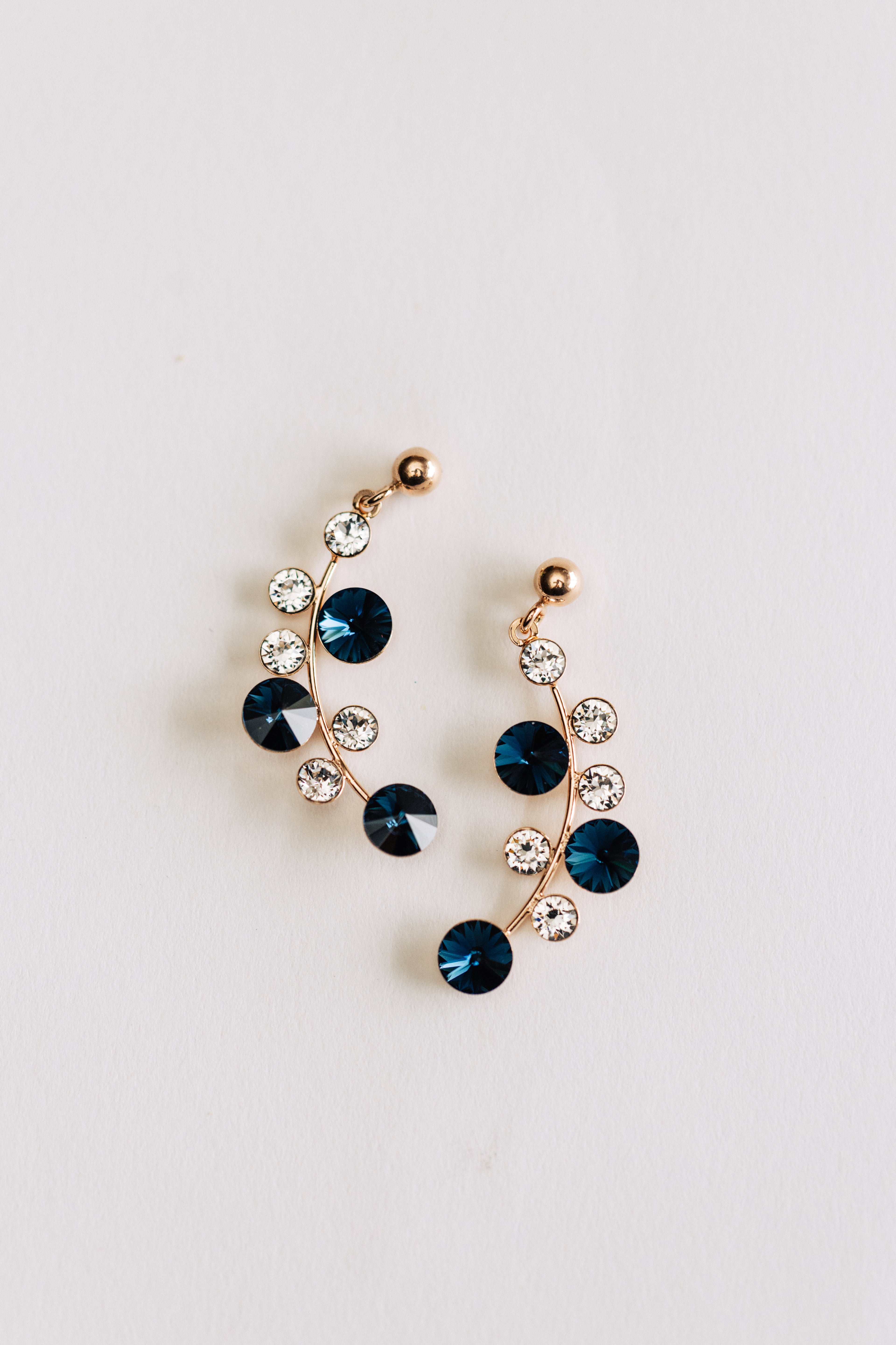 A pair of rose gold earrings featuring blue and clear crystals arranged in a circular pattern on a white background.