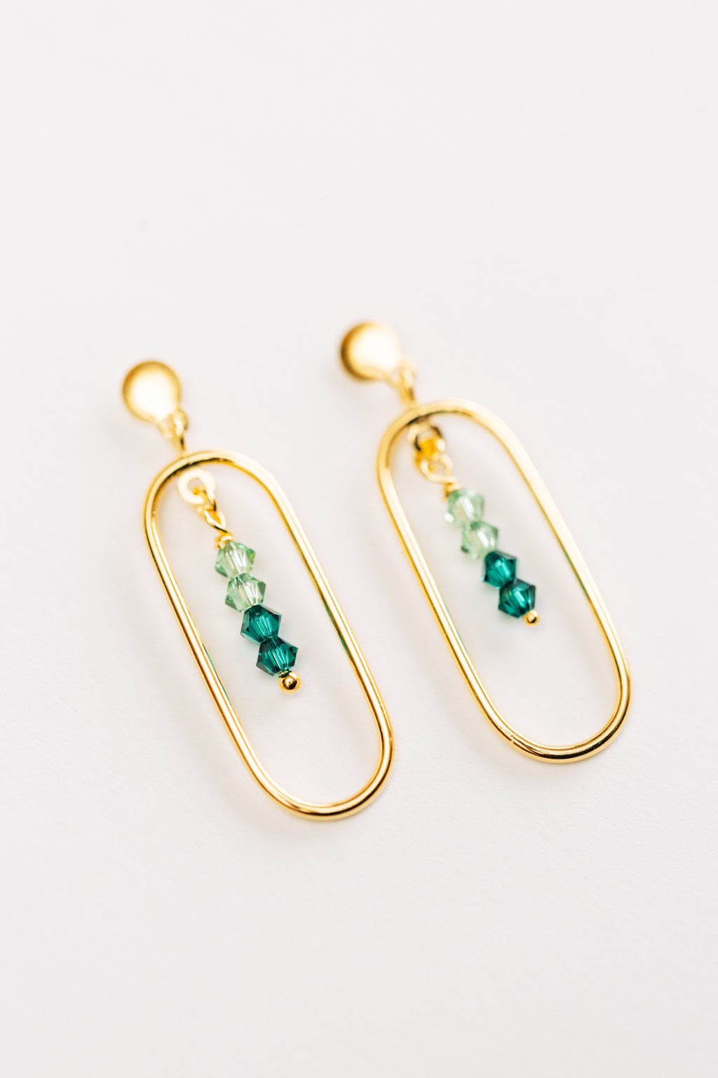 This image displays a pair of elegant hypoallergenic Adeline earrings, featuring a sophisticated gold color. The earrings are designed with a sleek, elongated oval frame that encases a series of small, gradient green crystals strung in a line.