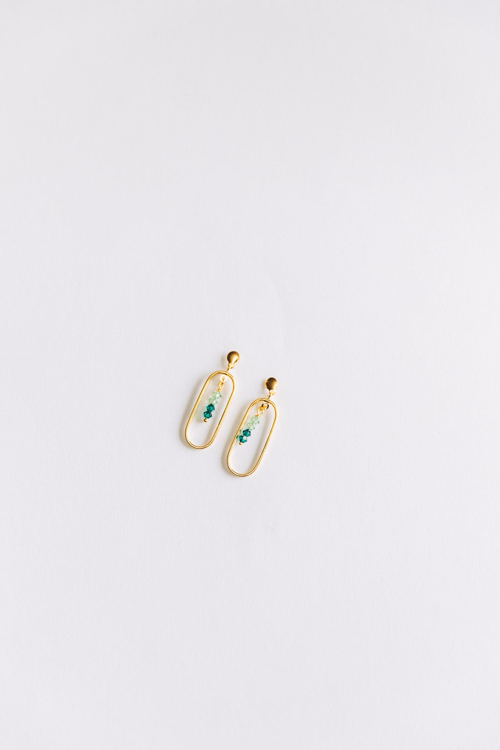 This image displays the Adeline earrings in gold, featuring a sleek and minimalist design. The earrings are composed of a small, elegant loop with a delicate green crystals at their center, adding a touch of color and sophistication.