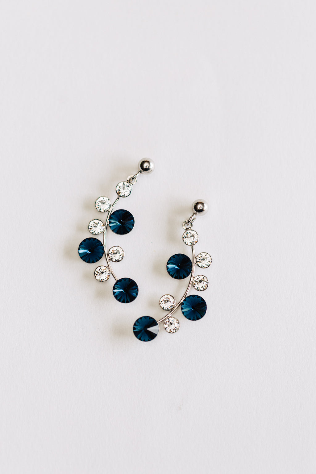 This image displays a pair of elegant hypoallergenic Aurora earrings in silver, featuring a captivating array of blue and clear crystals. The design incorporates a series of these crystals linked together, suspended from small, sleek silver hooks that enhance their delicate charm.