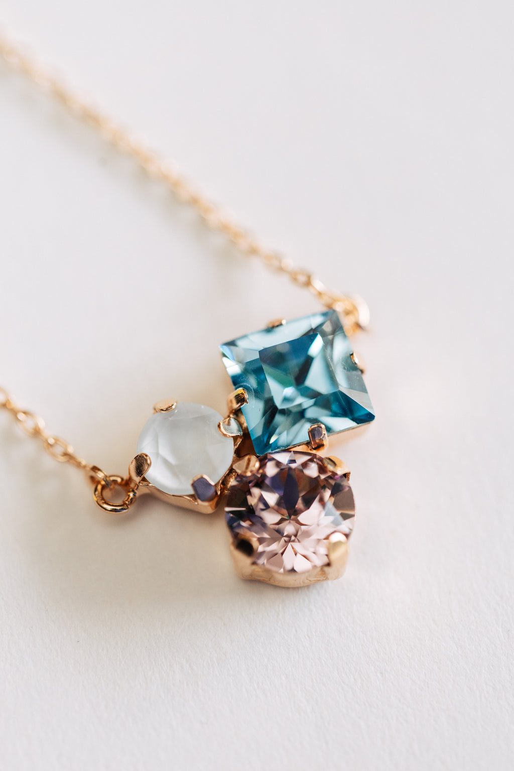 A hypoallergenic rose gold necklace with three stones pendants in blue, clear, and pink colors, attached to a gold chain.